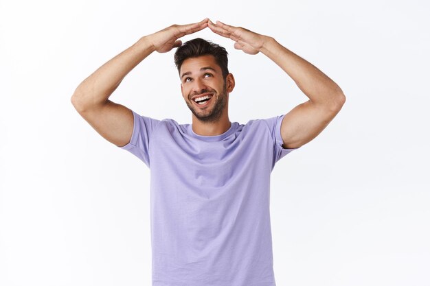 Chico sexy masculino barbudo atractivo alegre en camiseta púrpura en buena forma deportiva, levantar las manos por encima de la cabeza y mirar los brazos haciendo gesto de techo, casa, sonriendo encantado