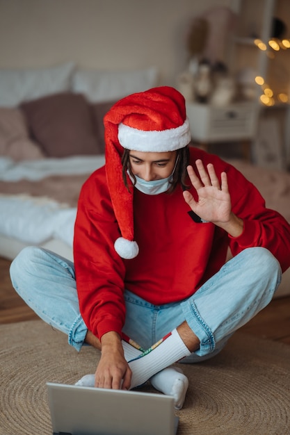 Chico con sombrero de santa cerca de la computadora portátil está interactuando a través de videollamadas. Navidad en aislamiento en casa. Distanciamiento social por vacaciones.