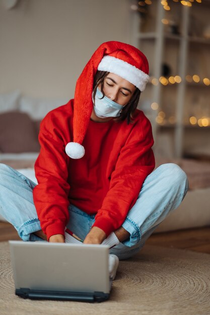 Chico con sombrero de santa cerca de la computadora portátil está interactuando a través de videollamadas. Navidad en aislamiento en casa. Distanciamiento social por vacaciones.