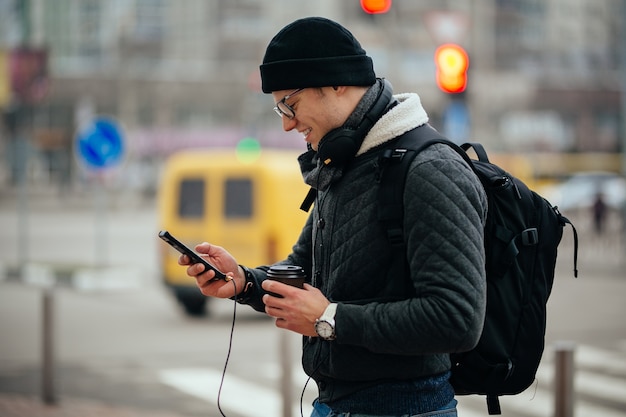 Chico sonriente en anteojos con auriculares, usando su teléfono inteligente, sosteniendo una taza de café caliente