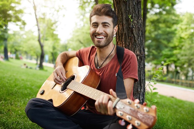 Chico sonriente atractivo sentado en el parque con guitarra, músico tocando y cantando