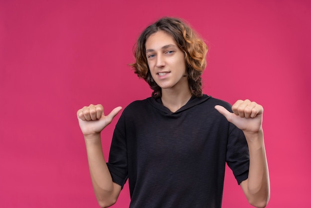 Foto gratuita chico sonriente con cabello largo en camiseta negra apunta a sí mismo en la pared rosa