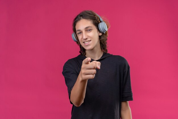 Chico sonriente con cabello largo en camiseta negra escucha música de auriculares y apunta hacia adelante en la pared rosa