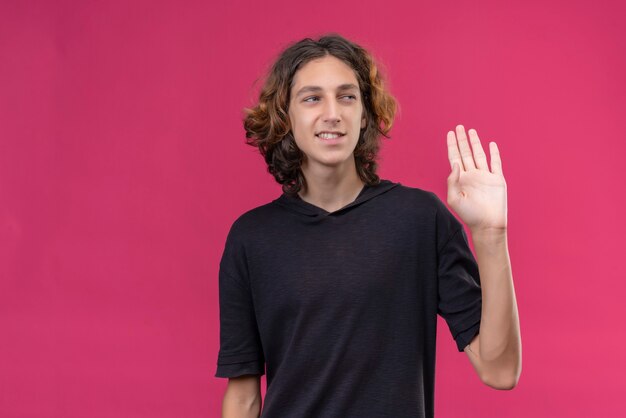 Chico sonriente con cabello largo en camiseta negra mostrando hola con su mano en la pared rosa