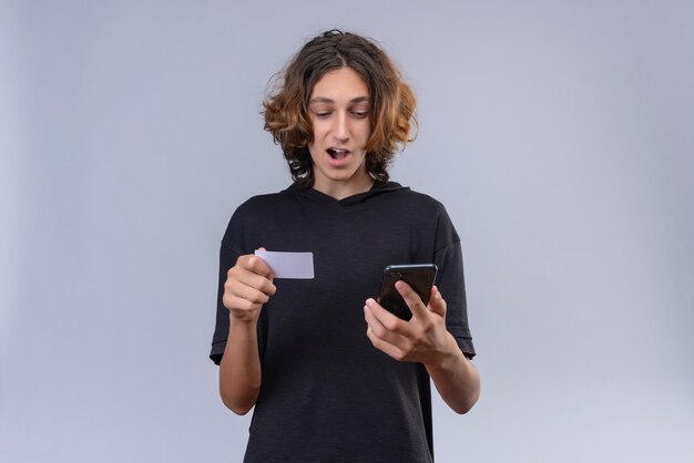 Chico sonriente con cabello largo en camiseta negra sosteniendo un teléfono y una tarjeta bancaria en la pared blanca