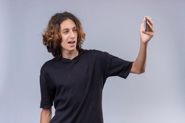 Chico sonriente con cabello largo en camiseta negra tomar un selfie en pared blanca