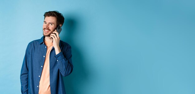 Chico sonriente moderno hablando por teléfono móvil mirando feliz a la cámara de pie sobre fondo azul.