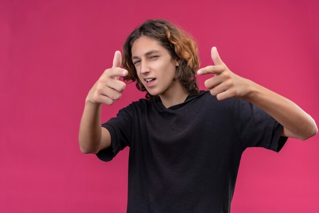 Chico sonriente con pelo largo en camiseta negra apunta hacia adelante en la pared rosa