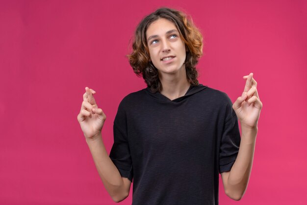 Chico sonriente con pelo largo en camiseta negra cruzando los dedos en la pared rosa