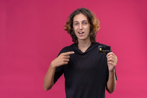 Chico sonriente con pelo largo en camiseta negra sosteniendo una tarjeta bancaria y apunta a la tarjeta en la pared rosa