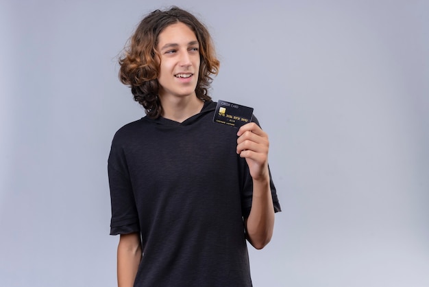 Foto gratuita chico sonriente con pelo largo en camiseta negra sosteniendo una tarjeta bancaria en la pared blanca