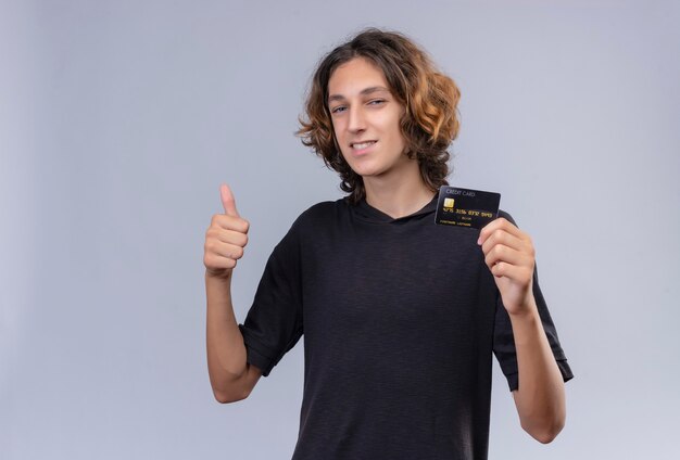 Chico sonriente con pelo largo en camiseta negra sosteniendo una tarjeta bancaria y pulgar levantado en la pared blanca