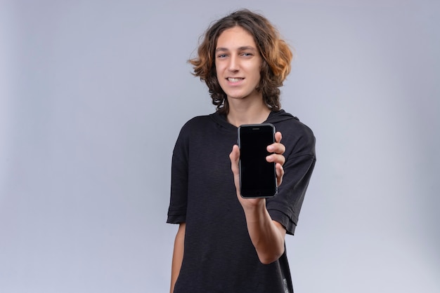 Chico sonriente con el pelo largo en camiseta negra sosteniendo un teléfono en la pared blanca