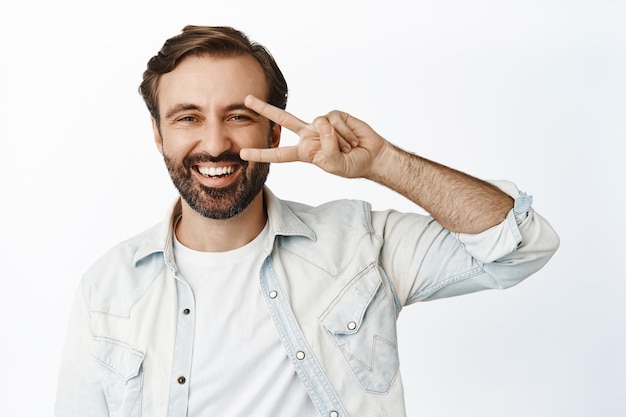 Chico sonriente positivo muestra signo de saludo de paz mirando feliz a la cámara de pie sobre fondo blanco.