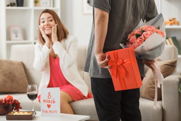 Chico sosteniendo presente con ramo mujer sonriente poniendo las manos en las mejillas sentado en el sofá en el día de la mujer feliz en la sala de estar