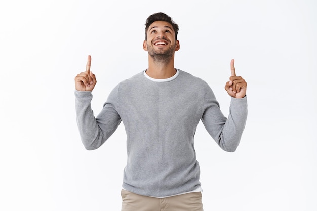 Foto gratuita chico con suéter gris sobre camiseta, levanta la cabeza y contempla la pancarta del producto interesante, apuntando hacia arriba intrigado