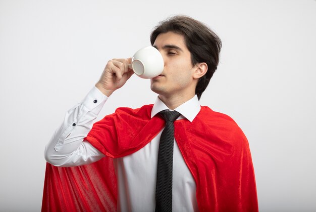 Chico superhéroe joven complacido con corbata bebe café de taza aislado sobre fondo blanco.