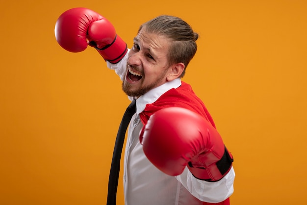 Chico superhéroe joven emocionado con corbata y guantes de boxeo de pie en pose de lucha aislado sobre fondo naranja