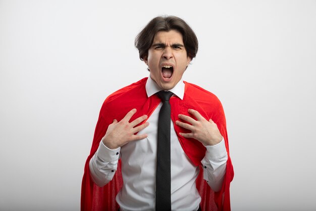 Chico superhéroe joven enojado mirando a cámara con corbata poniendo las manos en el pecho aislado en blanco