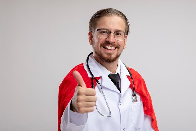 Chico superhéroe joven sonriente con bata médica con estetoscopio y gafas mostrando el pulgar hacia arriba