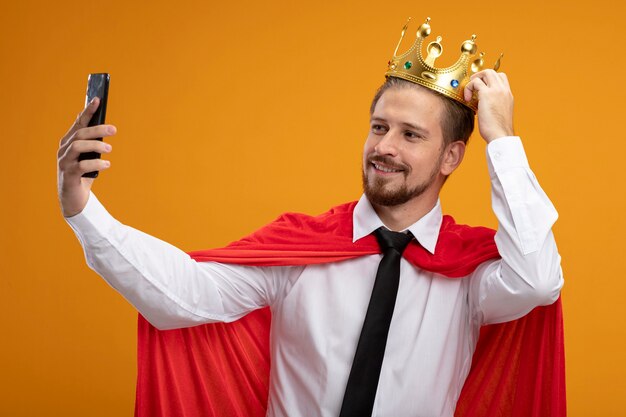 Chico superhéroe joven sonriente con corbata y corona poniendo la mano en la corona y tomar un selfie aislado sobre fondo naranja