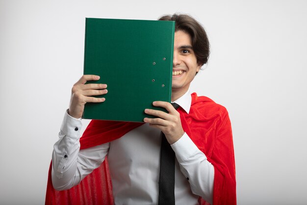 Chico superhéroe joven sonriente con corbata cubierta con el portapapeles aislado sobre fondo blanco.