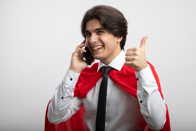 Chico superhéroe joven sonriente con corbata habla por teléfono y mostrando el pulgar hacia arriba aislado en blanco