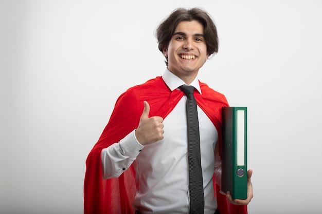 Chico superhéroe joven sonriente con corbata sosteniendo el portapapeles y mostrando el pulgar hacia arriba aislado en blanco