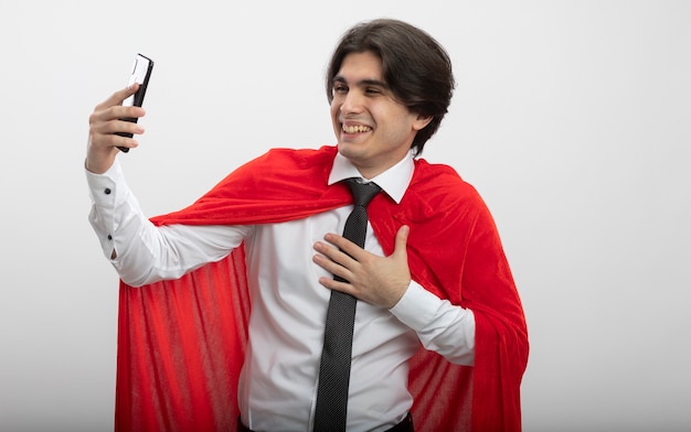 Chico superhéroe joven sonriente con corbata tomar un selfie y poner la mano en el pecho aislado sobre fondo blanco.