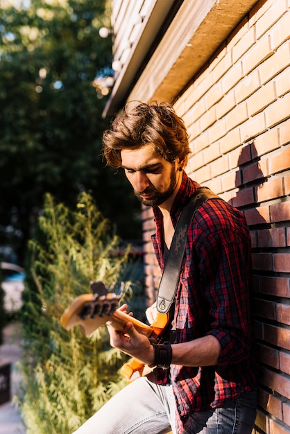 Foto gratuita chico tocando la guitarra eléctrica apoyándose en muro de ladrillo