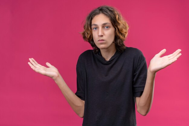 Chico triste con pelo largo en camiseta negra extendió sus manos en la pared rosa