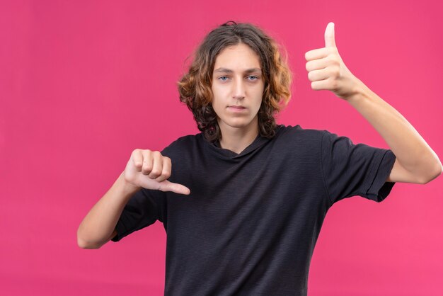 Chico triste con el pelo largo en camiseta negra mostrando el pulgar hacia arriba con una mano pulgar hacia abajo con la otra mano en la pared rosa