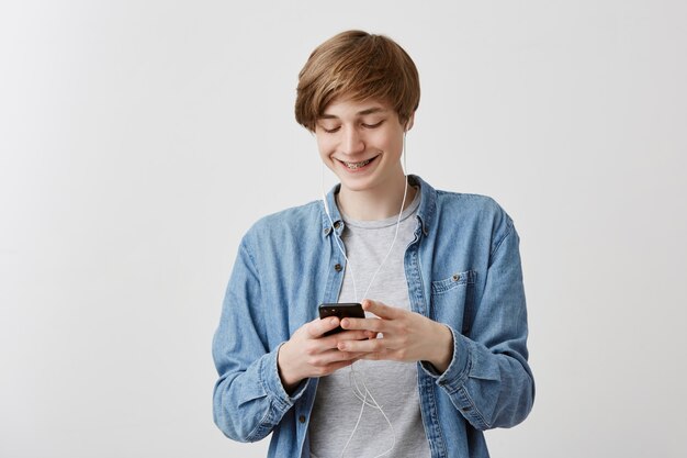 Chico de uacasian con el pelo rubio de pie contra el fondo gris que sostiene el teléfono inteligente descargando música usando la conexión a Internet con aspecto complacido, emocionado, sonriendo mientras mira la pantalla del teléfono celular