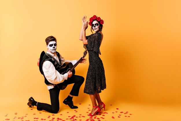 Chico zombi cantando una serenata para su novia muerta. Retrato de interior de pareja espeluznante celebrando halloween juntos.