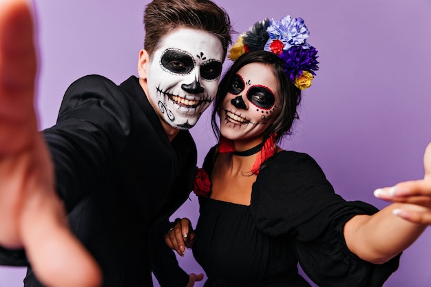 Chico zombie positivo haciendo selfie en estudio con novia. Amigos felices en disfraces de disfraces divirtiéndose.