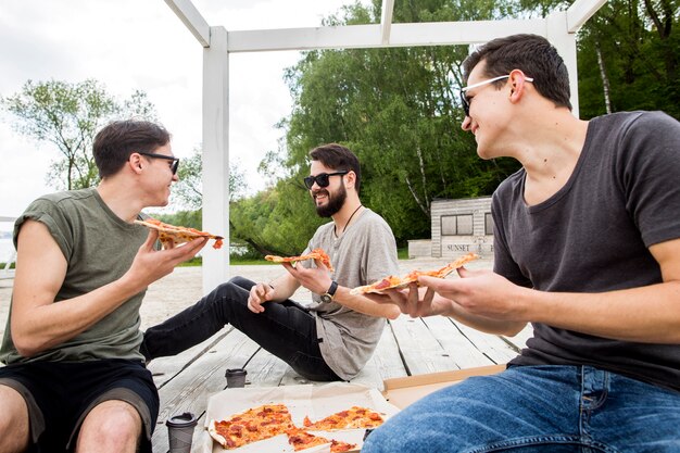 Chicos jóvenes con trozos de pizza conversando en la playa