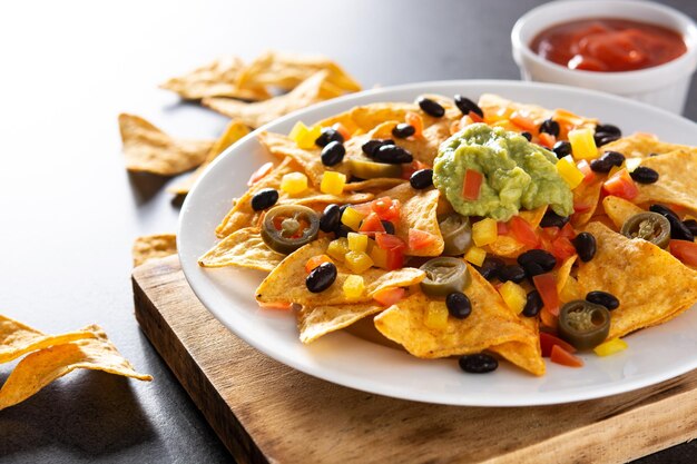 Chips de tortilla de nachos mexicanos con frijoles negros, guacamole, tomate y jalapeño sobre fondo negro