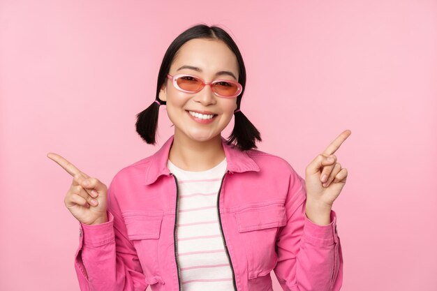 Choice Elegante chica coreana modelo femenino asiático señala con el dedo hacia los lados muestra dos variantes de anuncios de productos que muestran artículos de pie sobre fondo rosa