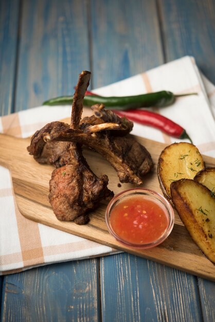 Chuletas de cordero de alto ángulo sobre tabla de madera con papas y salsa