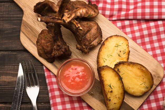 Chuletas de cordero planas sobre tabla de madera con papas y salsa