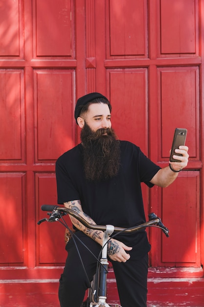 Ciclista joven sonriente que toma el selfie en su smartphone delante de puertas rojas