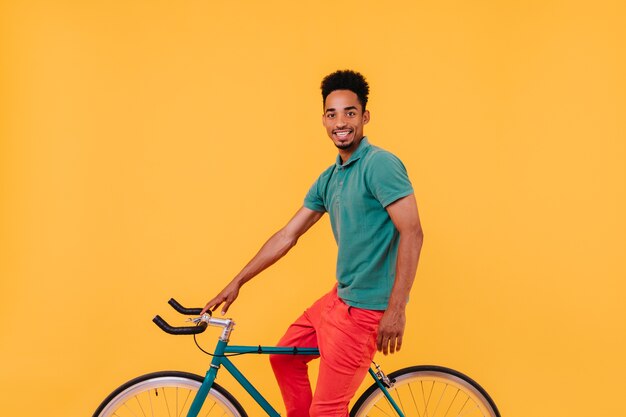 Ciclista masculino refinado posando con una sonrisa. Chico morena interesado sentado en bicicleta junto a la pared amarilla.