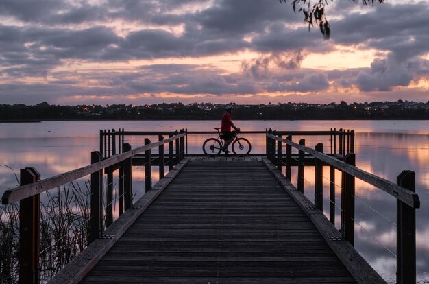 Ciclista de pie sobre un muelle de madera en el agua bajo un cielo nublado durante la puesta de sol por la noche
