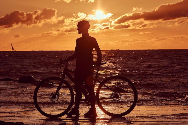 Ciclista de pie con su bicicleta y disfrutando de la puesta de sol en la costa del mar.