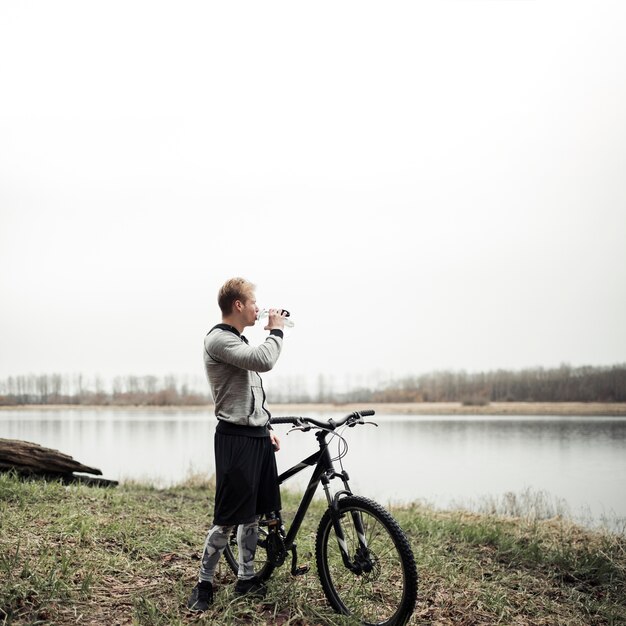 Ciclista que se coloca con la bicicleta que mira el lago mientras que bebe el agua de la botella