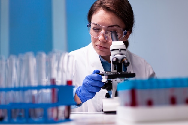 Científica en bata blanca mirando en microscopio de gama alta para experiencia en farmacia
