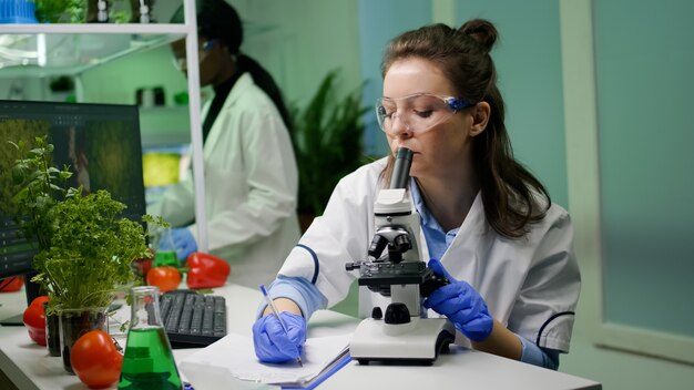 Científico biólogo mirando la muestra de hoja con microscopio médico mientras escribe experiencia en OMG en el bloc de notas. Químico examina el descubrimiento biológico en la planta que trabaja en el laboratorio farmacéutico