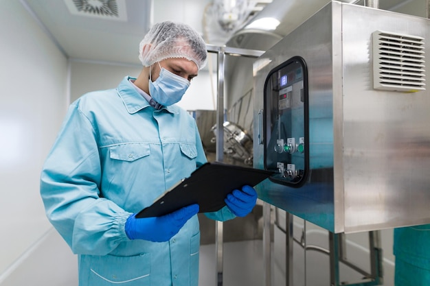 Científico caucásico con uniforme de laboratorio azul parado cerca de una gran máquina de acero con panel de control toma notas sobre la máquina