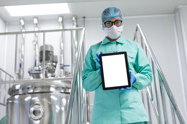 Científico caucásico con uniforme de laboratorio azul parado en escaleras cromadas y sosteniendo una tableta vacía hacia la cámara