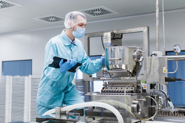 Un científico caucásico con uniforme de laboratorio azul toma notas sobre una gran máquina de acero cromado